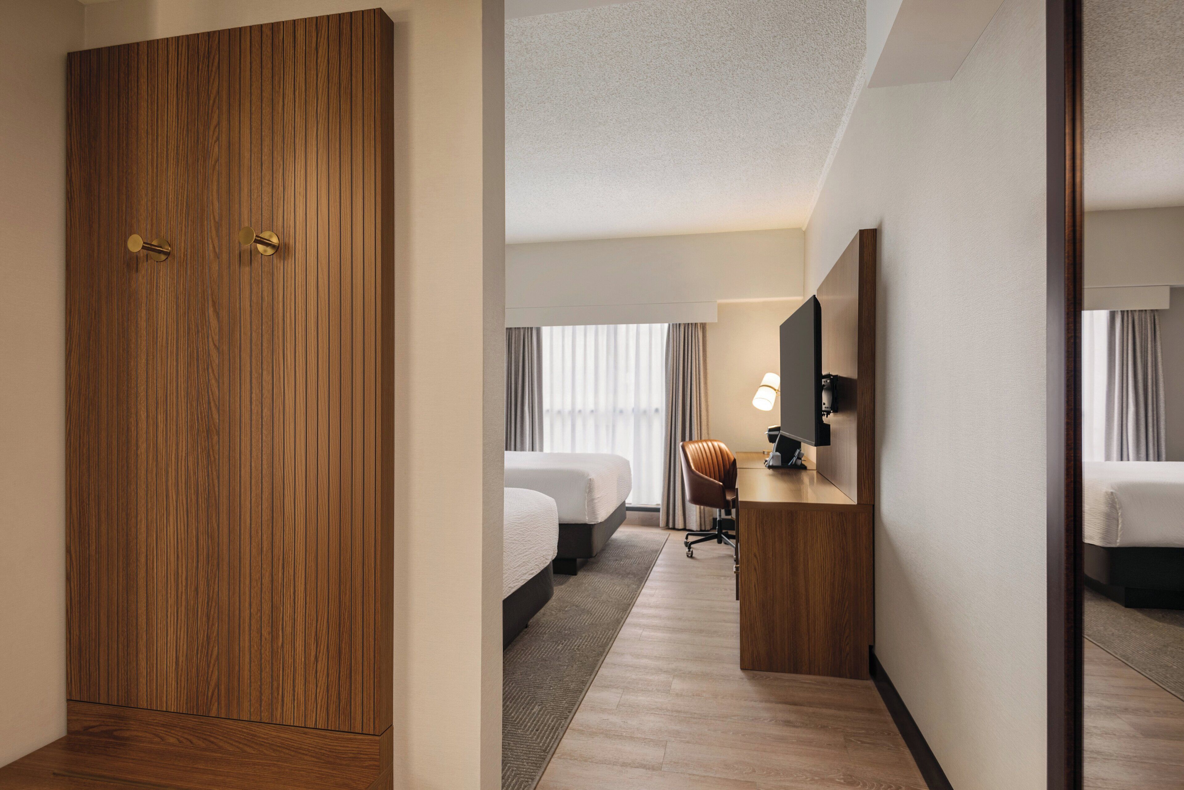 Een hotelkamer met een houten garderobe, een bureau met een lamp, twee bedden en een grote spiegel.