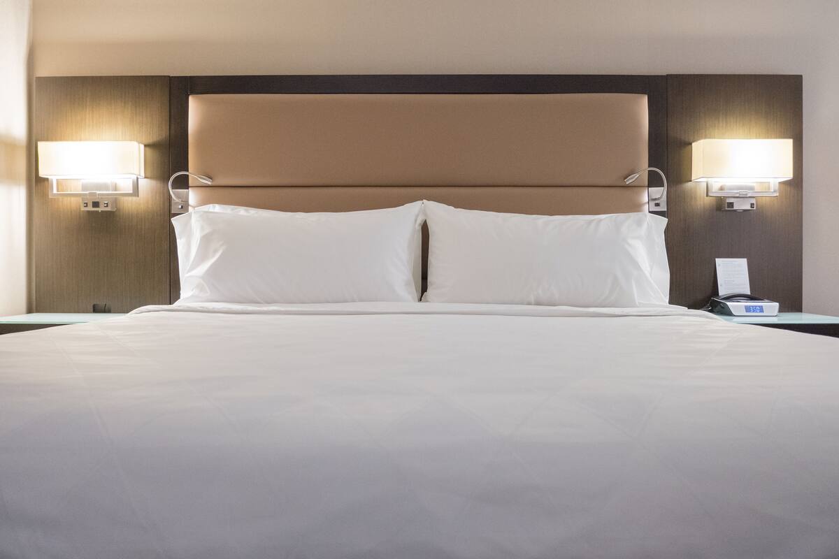 Quarto de hotel com uma cama grande, duas luminárias de cabeceira e mesa de apoio com telefone e abajur.