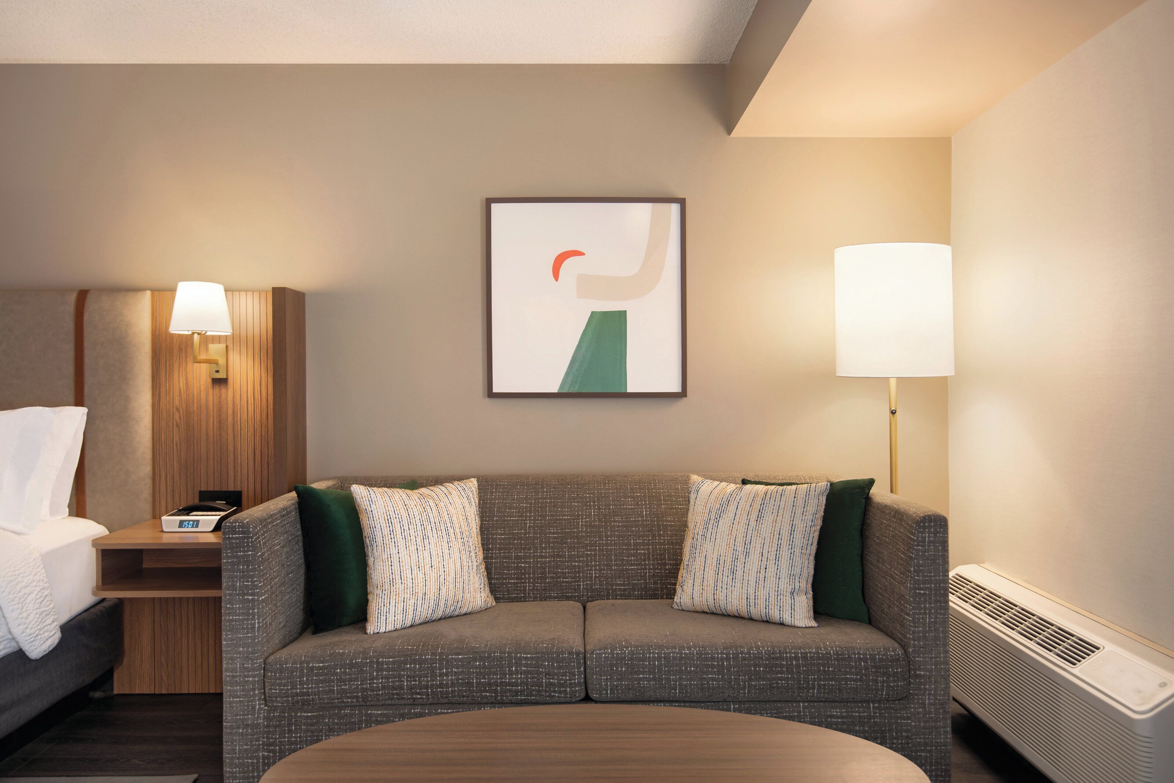 A hotel room with a gray sofa, two pillows, a framed abstract artwork, a bedside table with a lamp, and a heater.