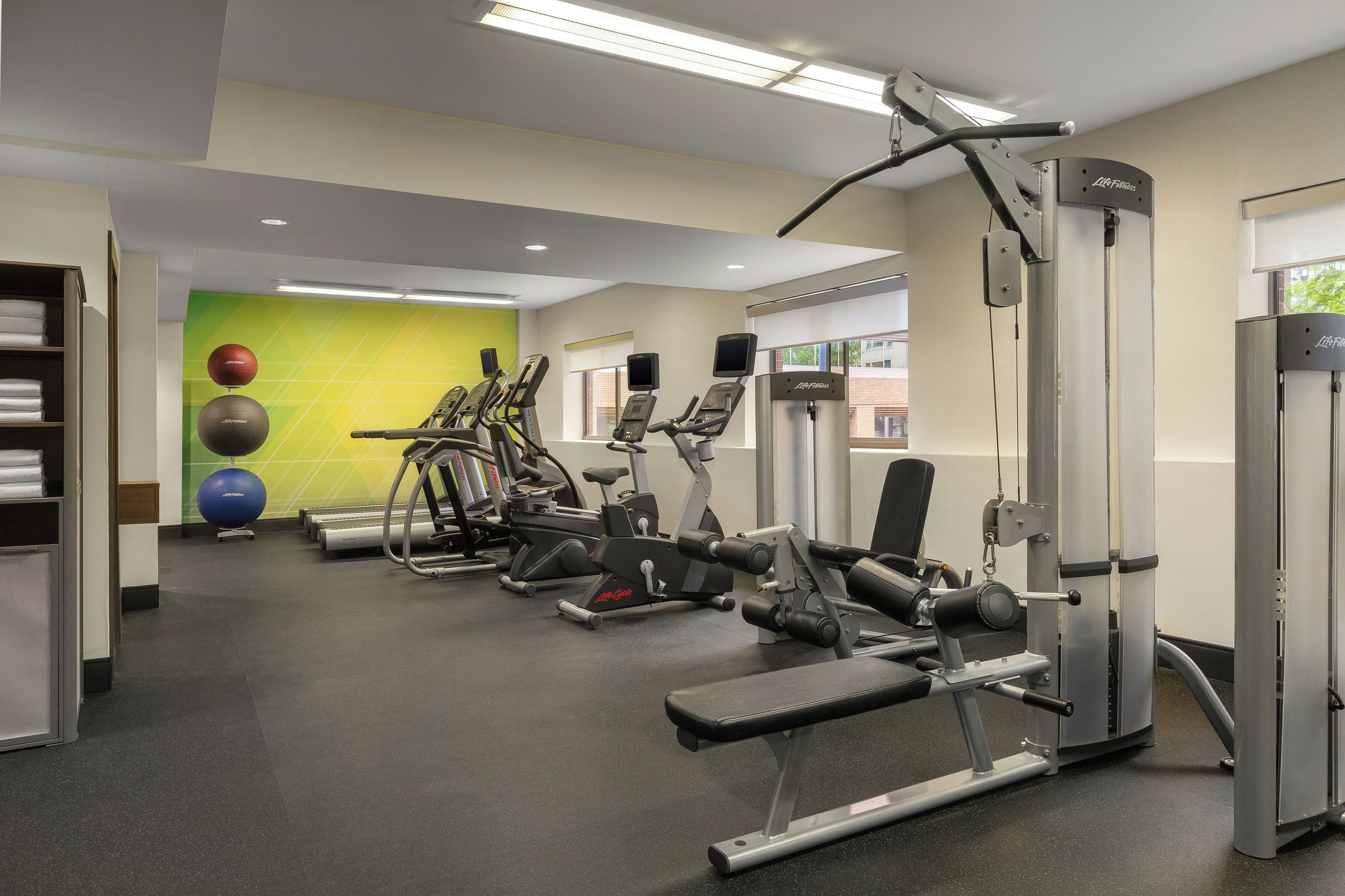 Een sportschool met diverse fitnessapparaten, een muur met oefenballen en een raam dat voor daglicht zorgt.