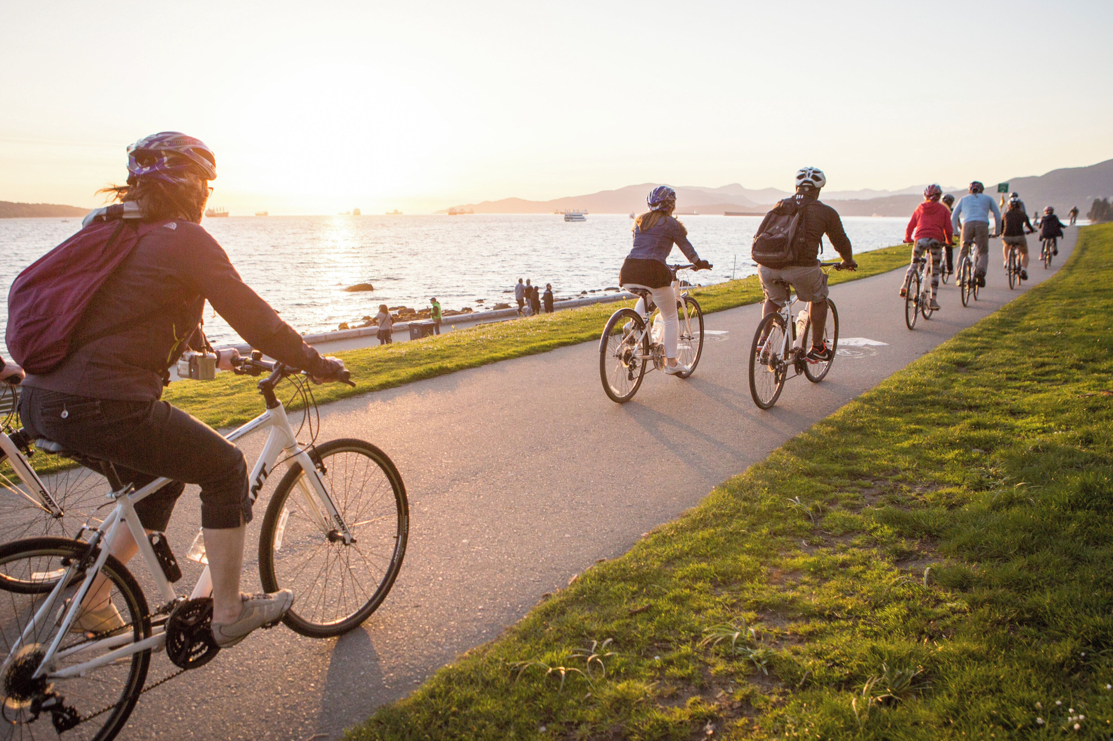 A group of cyclists on a paved path by a body of water with a sunset in the background.