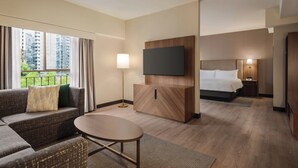 A modern hotel room with a sofa, a small round table, a TV mounted on a wooden cabinet, a bed with a headboard, and a nightstand with a lamp. - Holiday Inn & Suites Vancouver Downtown by IHG (Vancouver)