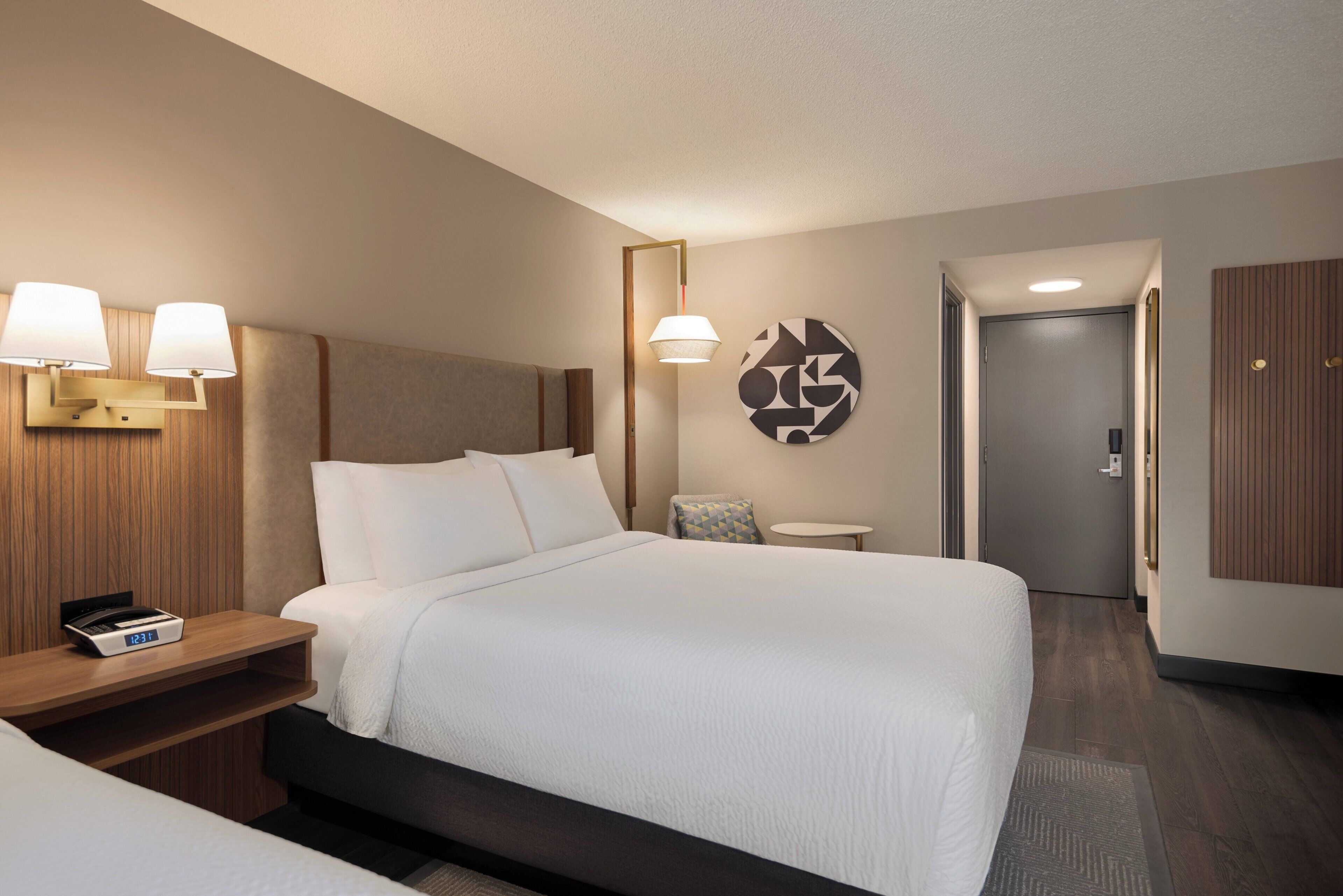 A hotel room with a bed, bedside tables, a clock, a lamp, a wall art piece, and a door.