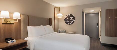 A hotel room with a bed, bedside tables, a clock, a lamp, a wall art piece, and a door.