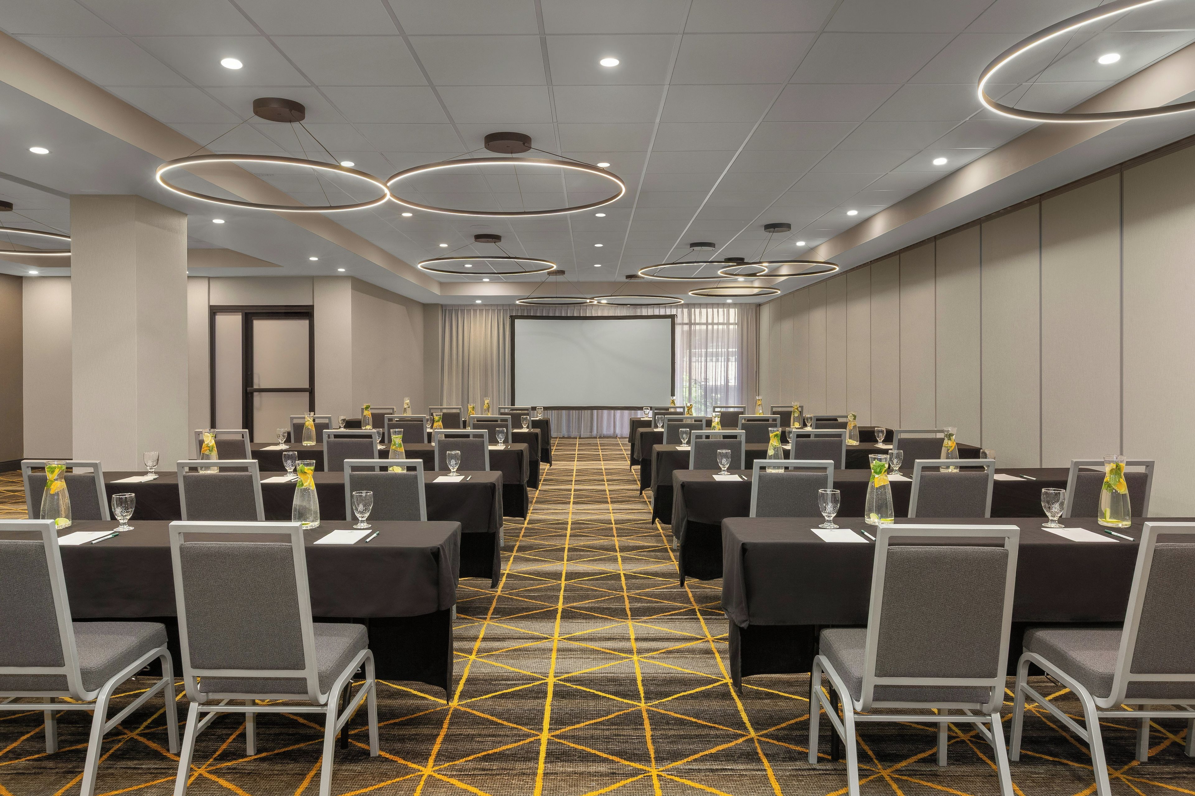 A conference room with rows of tables and chairs, a projector screen, and a patterned carpet.