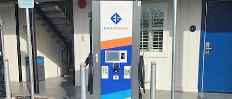 Una estación de carga para vehículos eléctricos EVGATEWAY con un panel de control, ubicada frente a un edificio con una puerta azul y una escalera.