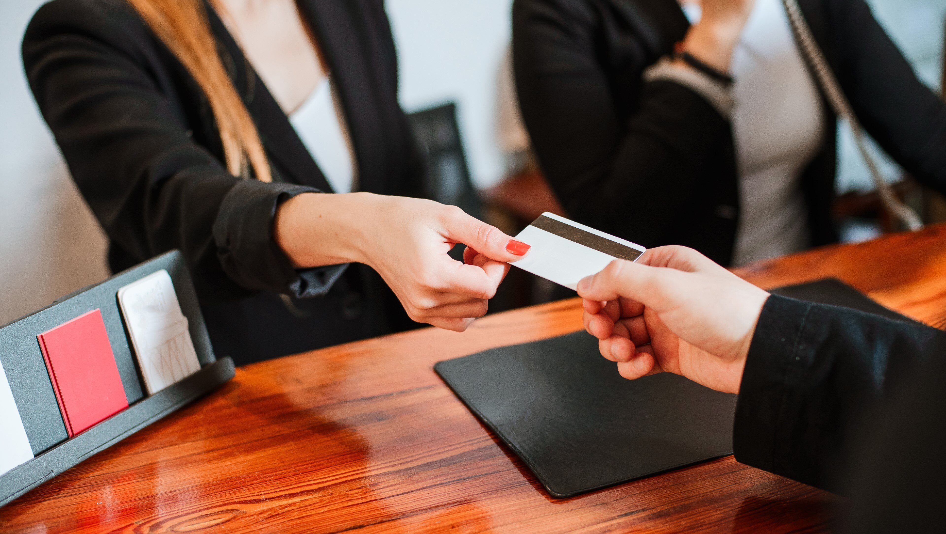 A person handing over a credit card to another person at a wooden table with a black folder and a red card on it.