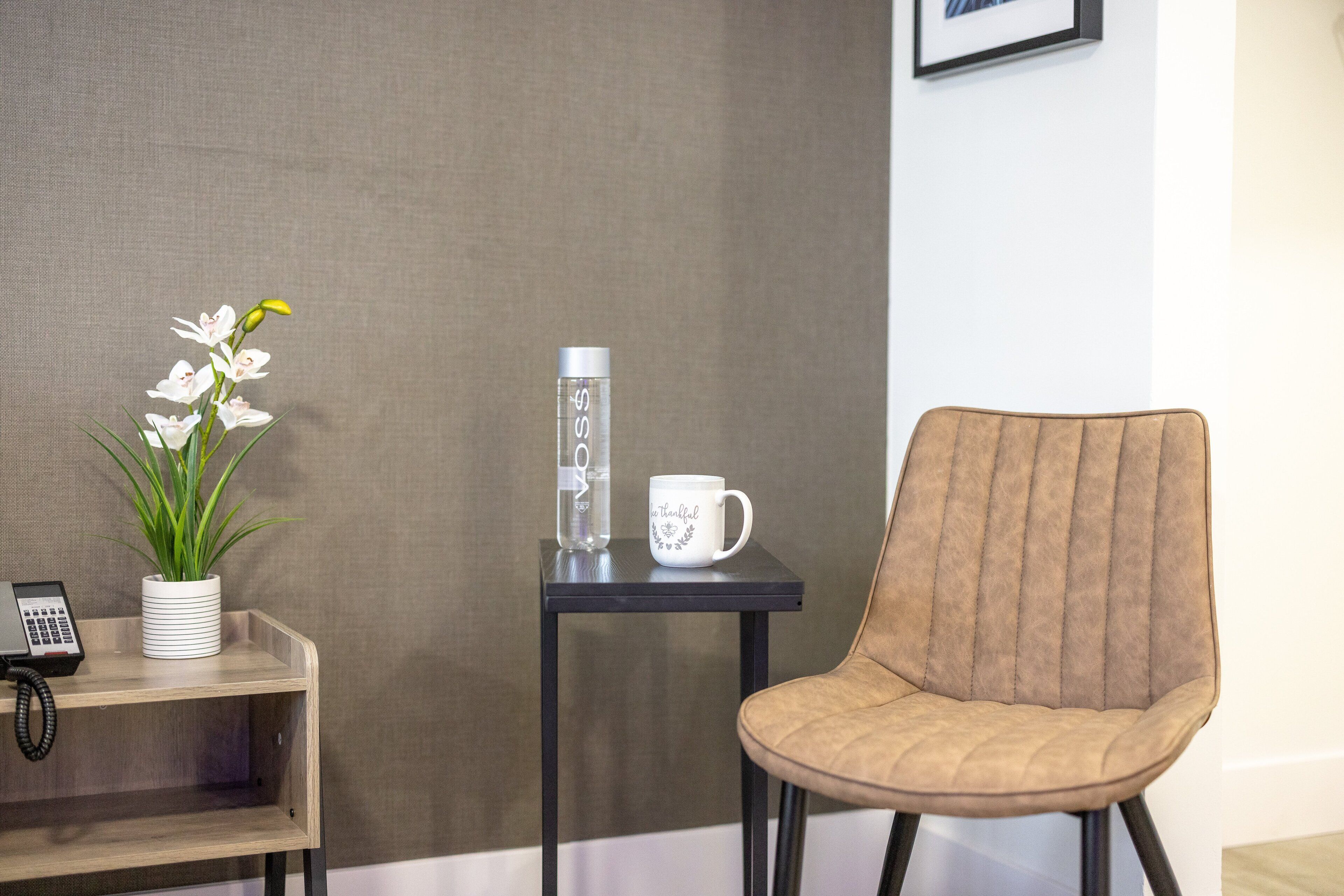 A room with a wooden shelf holding a potted plant and a telephone, a small black table with a water bottle and a white mug, and a brown upholstered chair.