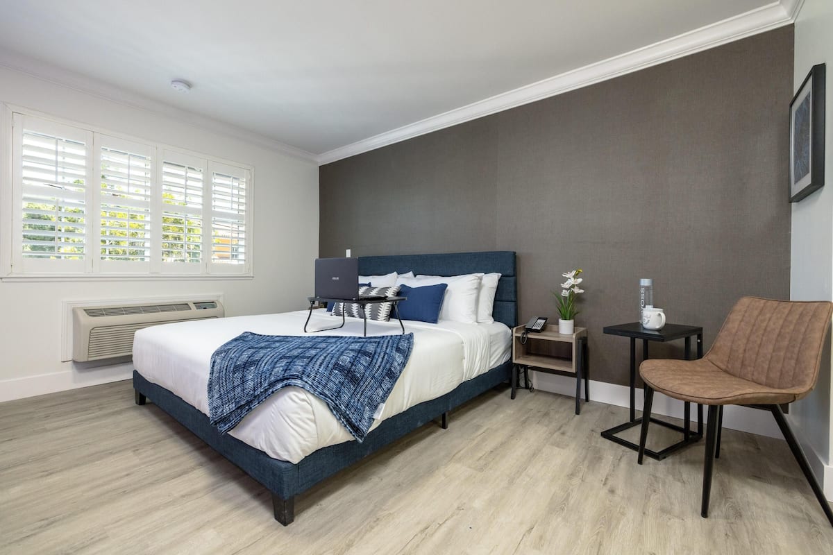 A bedroom with a bed, a chair, a nightstand, a lamp, and a window with blinds.