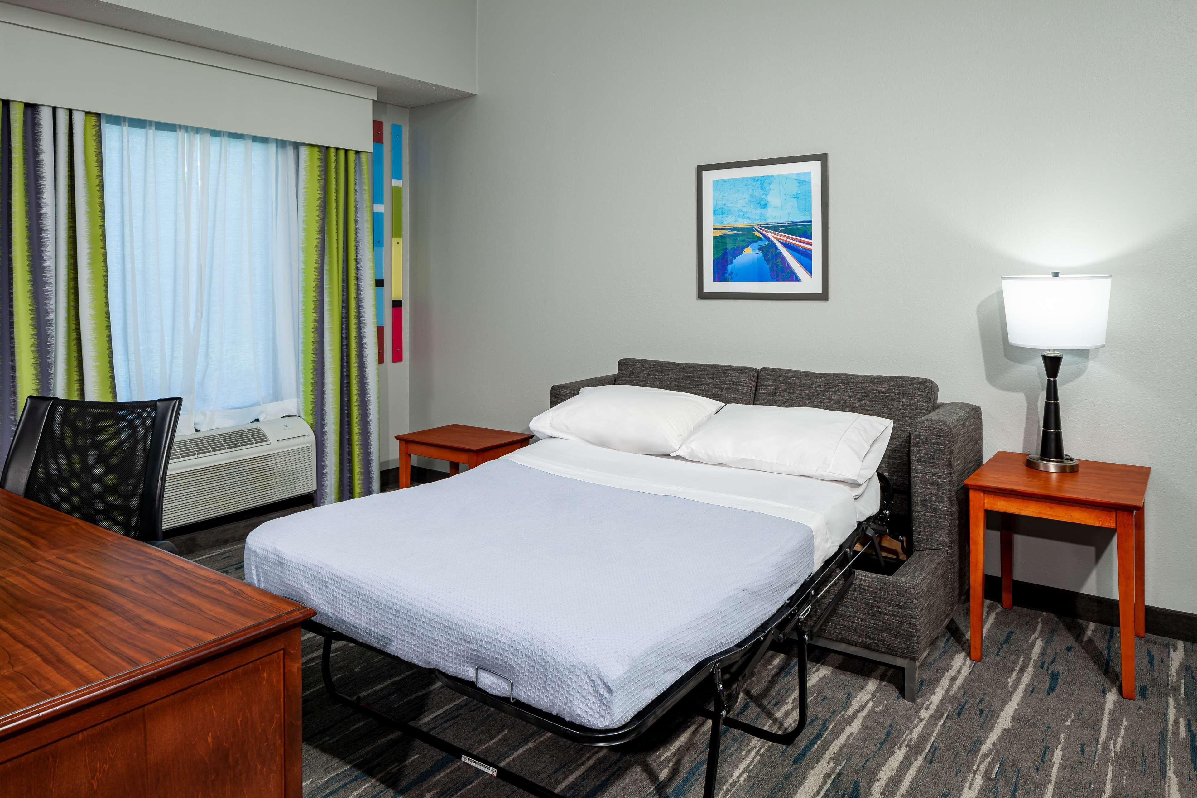 A hotel room with a bed, a desk, a chair, a lamp, and a picture on the wall.