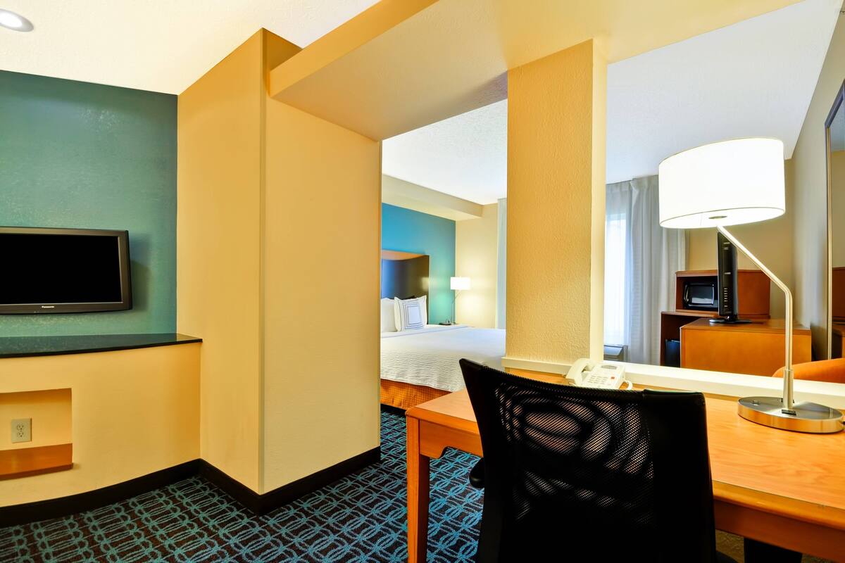 A hotel room with a flat-screen TV, a desk with a lamp, a chair, and a bed with a blue headboard.