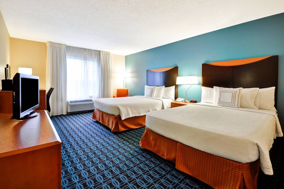 A hotel room with two beds, a desk with a TV, a lamp, and a window with curtains.