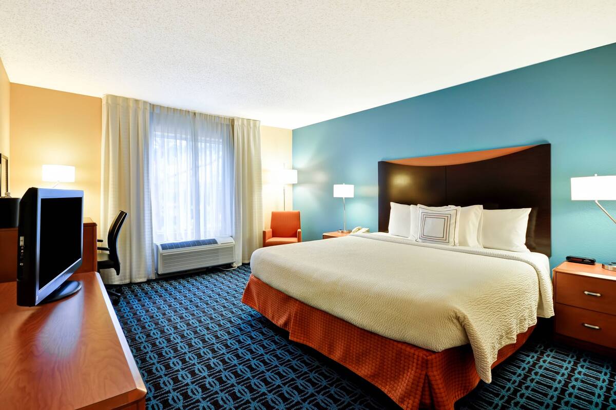A hotel room with a large bed, a desk with a computer, a chair, a window with curtains, and a wall-mounted TV.