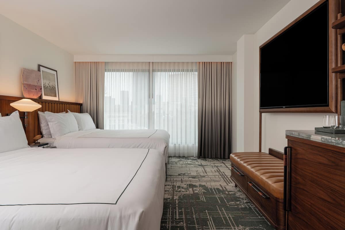 Habitación de hotel con dos camas, televisor, escritorio y un ventanal grande con cortinas.