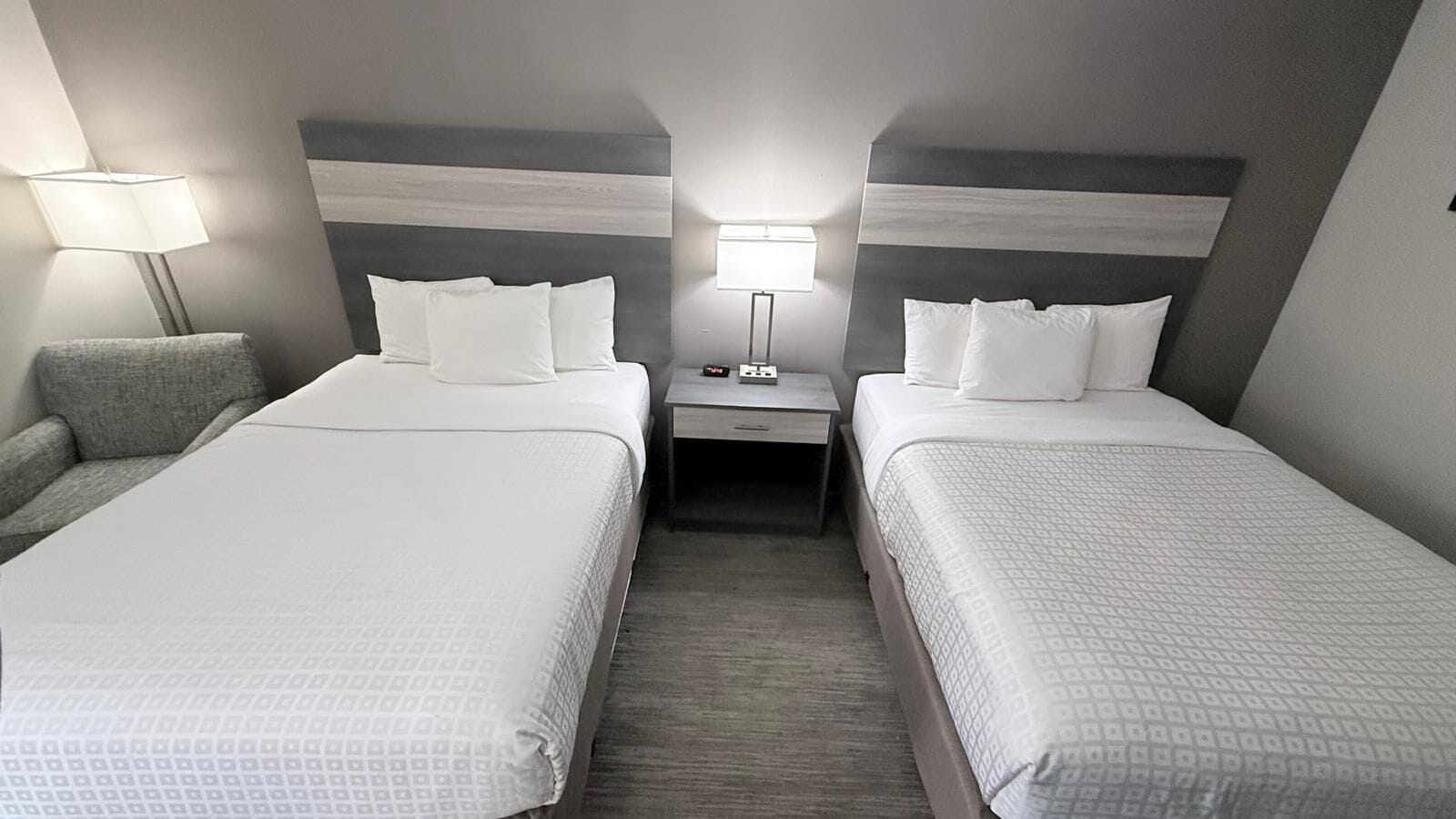 A hotel room with two beds, a nightstand, a lamp, and a chair.