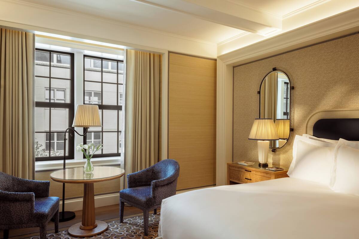 A hotel room with a large bed, two armchairs, a small round table with a lamp, a mirror, and a window with curtains.