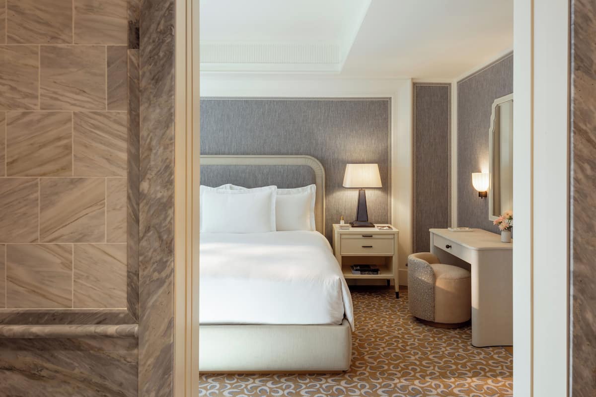 A hotel room with a large bed, a bedside table with a lamp, a vanity with a mirror, and a patterned floor.