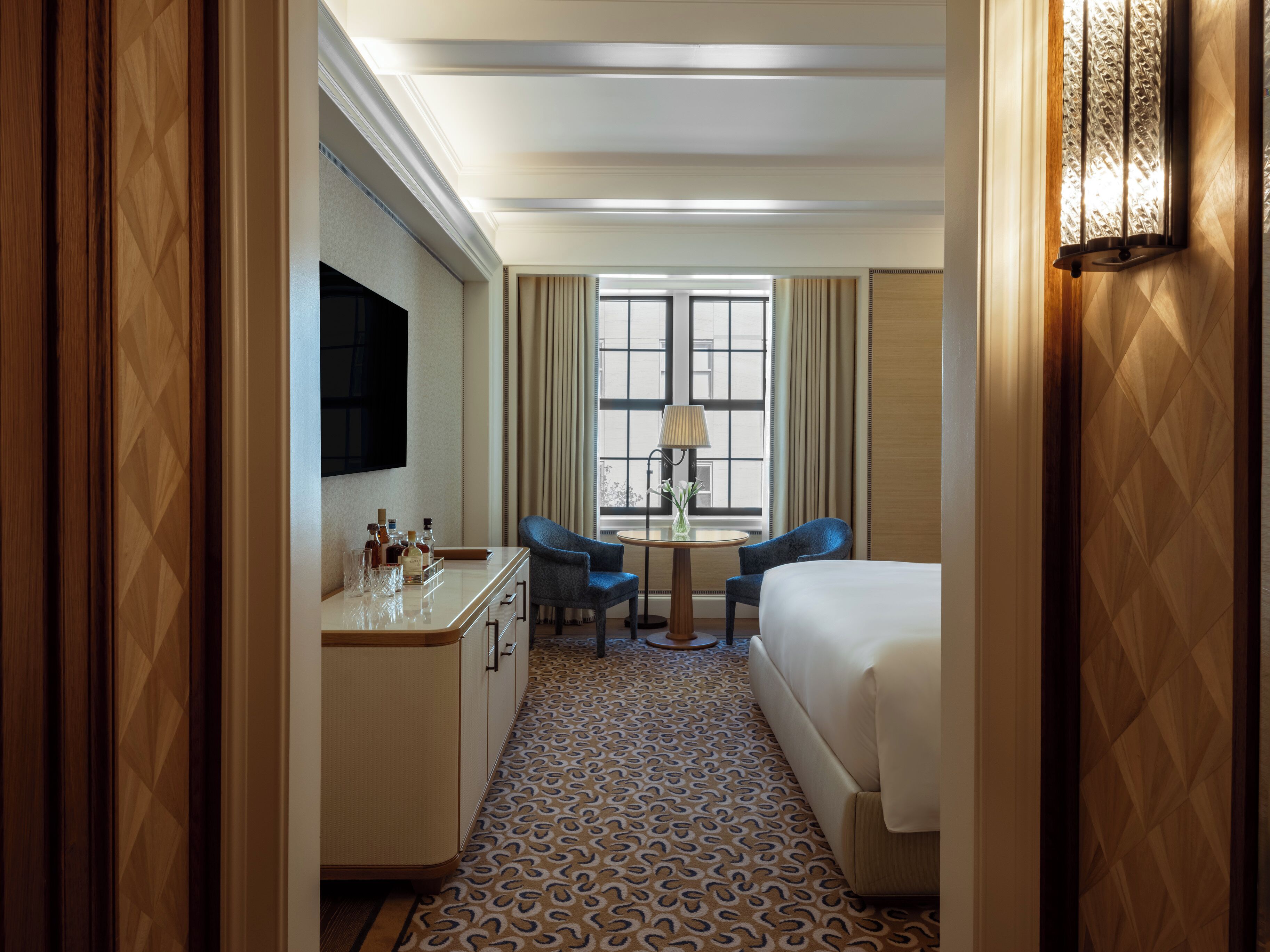 A hotel room with a bed, a desk with a lamp, two chairs, a TV, and a patterned carpet.