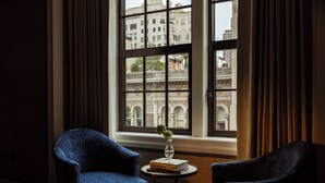 A room with two armchairs, a small table with a book and a vase, and a large window with a view of a building. - The Surrey A Corinthia Hotel (New York)