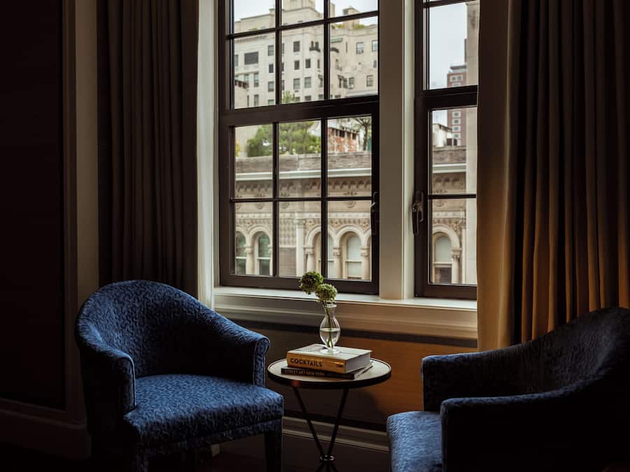 A room with two armchairs, a small table with a book and a vase, and a large window with a view of a building.