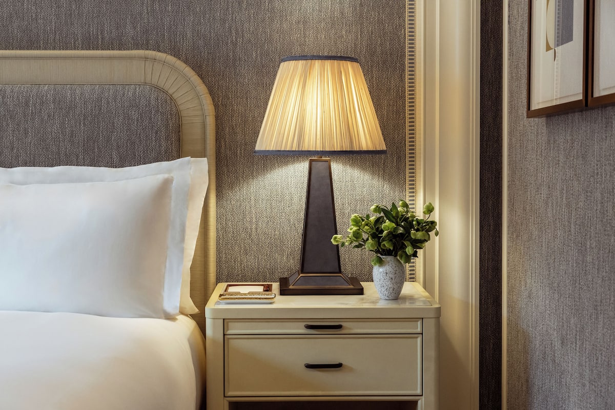 A neatly made bed with white linens, a bedside table with a lamp, a small vase with green plants, and a framed picture on the wall.
