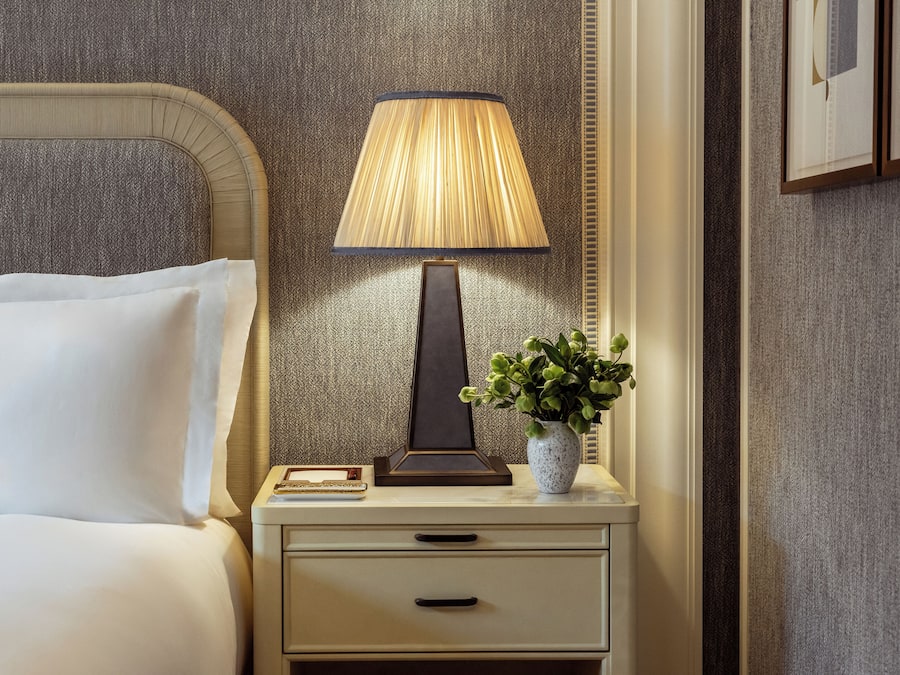 Un lit bien fait, avec du linge de lit blanc, une table de chevet avec une lampe, un petit vase contenant des plantes vertes et un tableau encadré au mur.