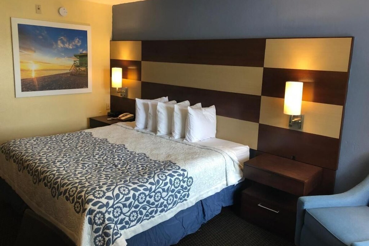 Une chambre d’hôtel comprenant un lit, des tables de chevet, une petite table, une chaise, un tableau au mur et une lampe fixée au mur.
