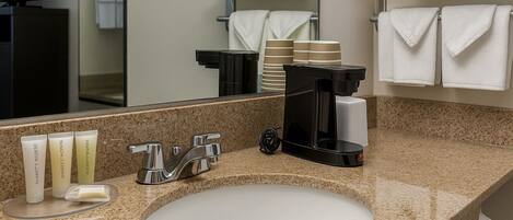 A hotel bathroom with a granite countertop, a white sink, a coffee maker, and toiletries.