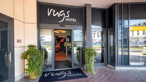 De ingang van Rugs Hotel, met een modern ontwerp, glazen deuren en een zwarte deurmat.