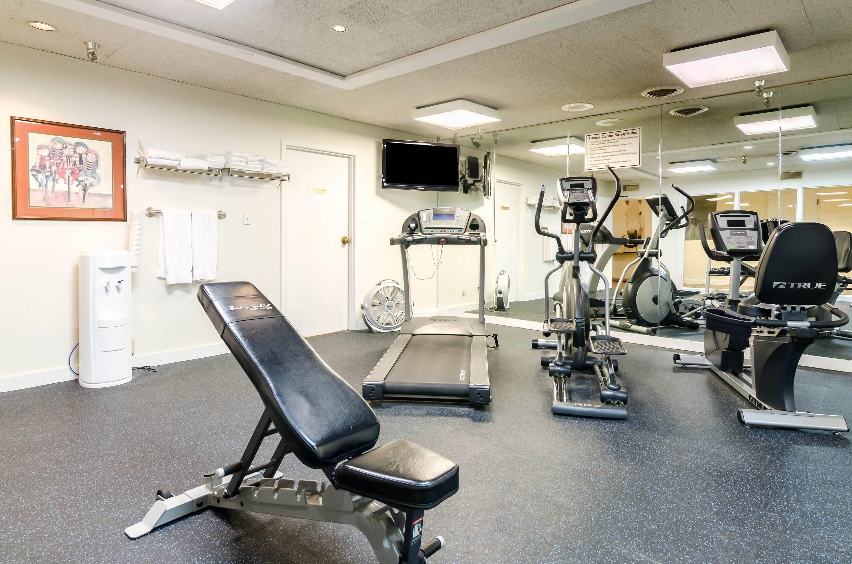 Een sportschool met een bankje, loopband en diverse fitnessapparaten.