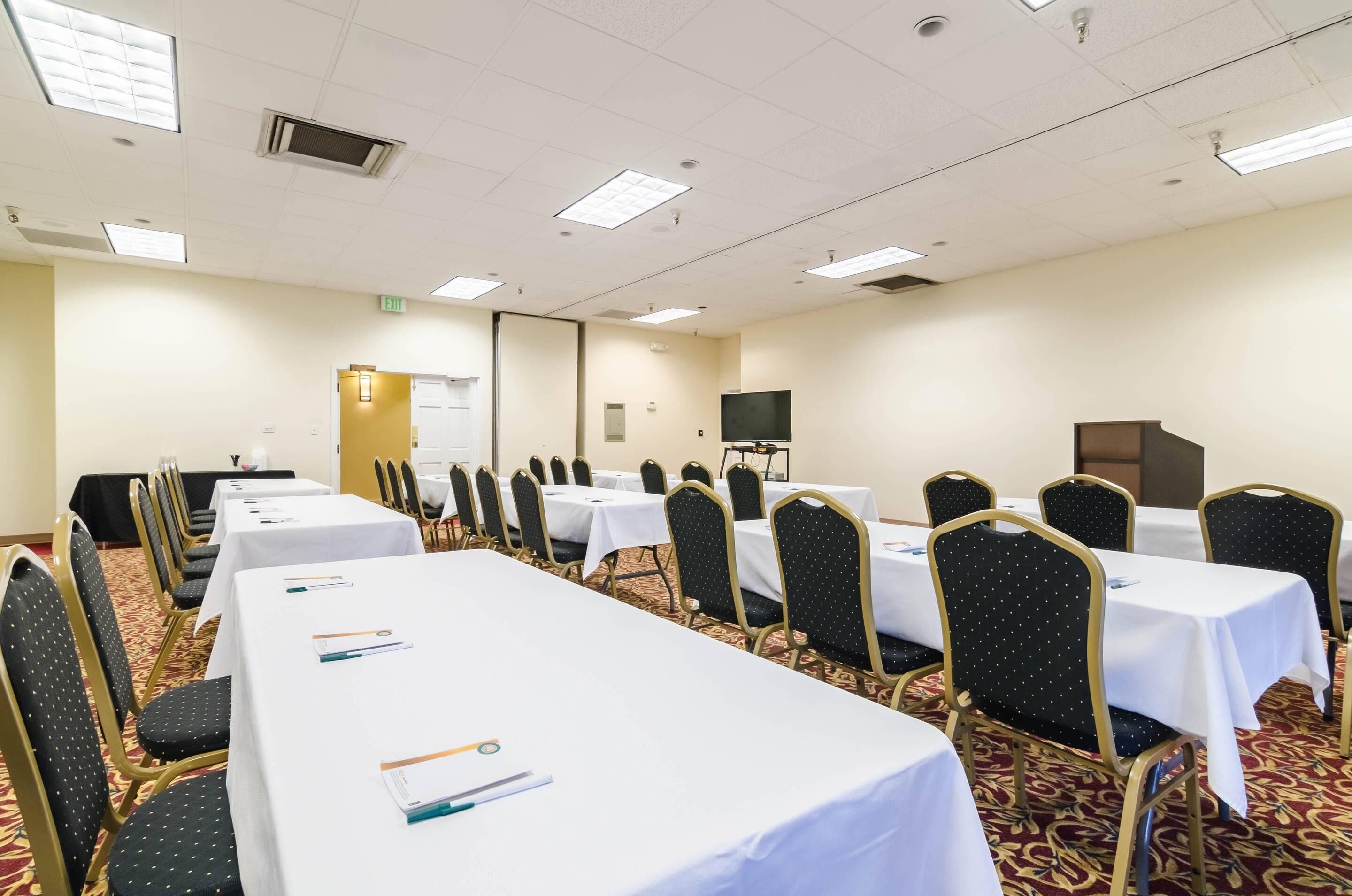 Un salón de conferencias con mesas y sillas dispuestas en filas, una pantalla de proyector y una pizarra.