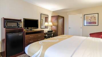 A hotel room with a bed, a desk with a computer, a microwave, a refrigerator, a TV, and a door.