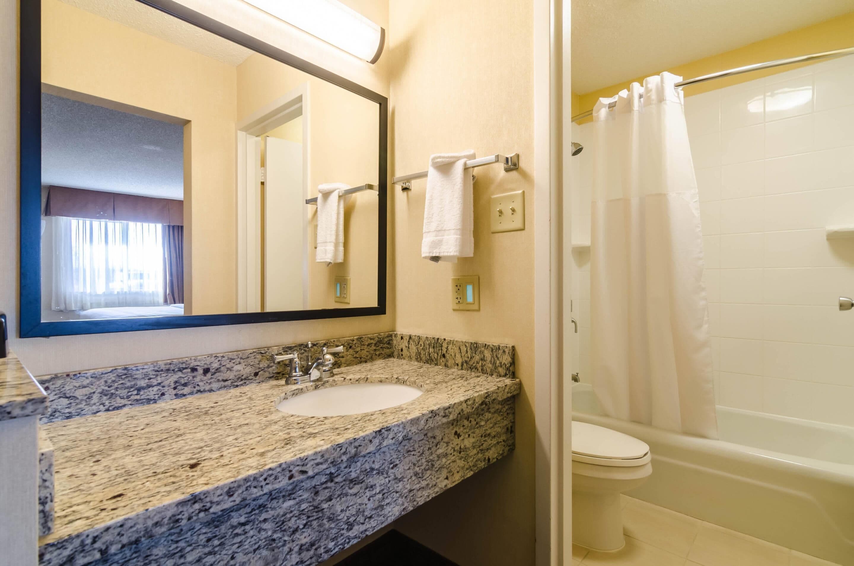 A bathroom with a granite countertop sink, a mirror, a towel rack, a toilet, and a bathtub with a shower curtain.