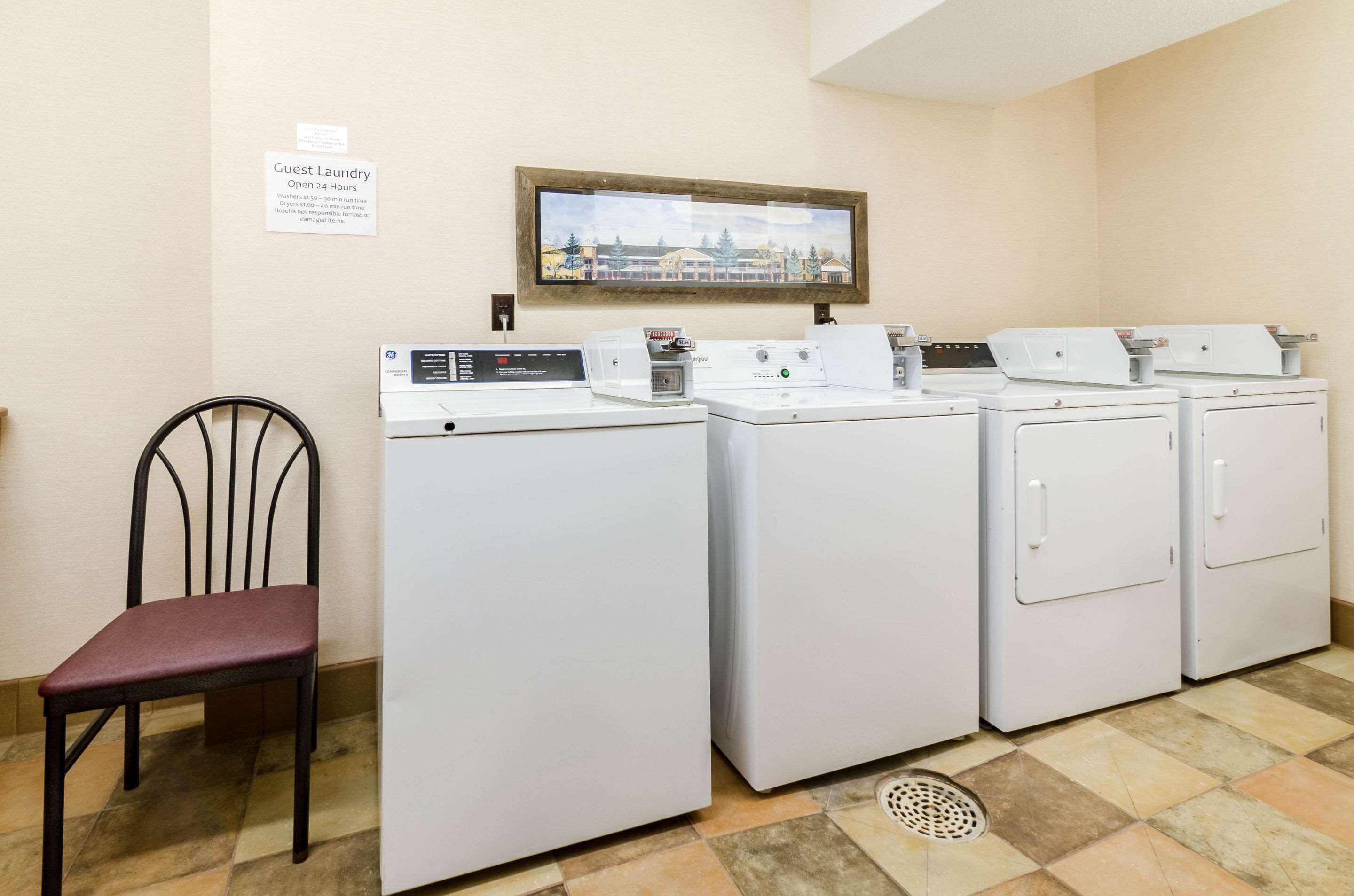 A laundry area with four washing machines, a chair, and a sign indicating guest laundry is open 24 hours.
