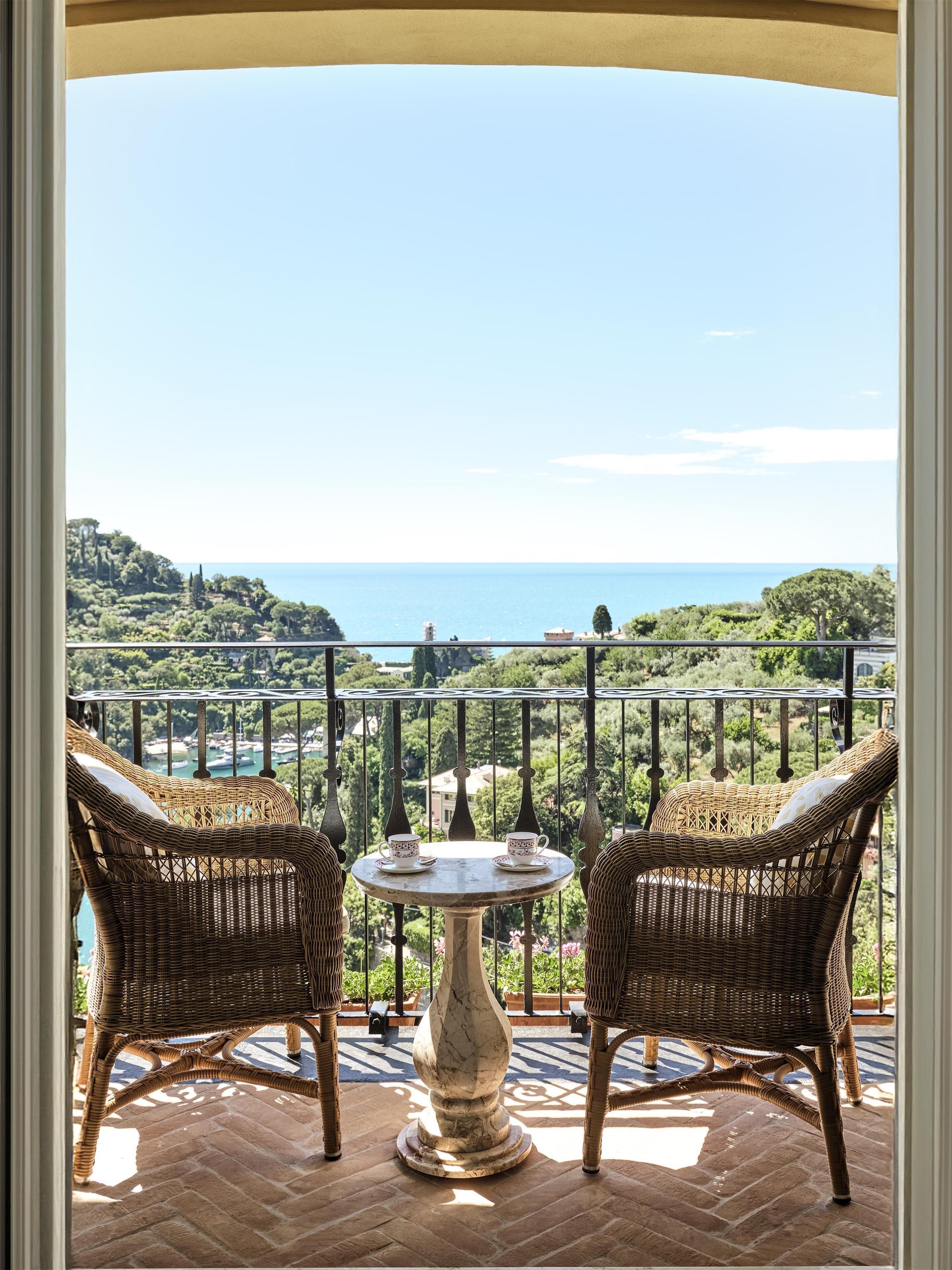 A balcony with wicker chairs and a table, overlooking a scenic landscape with trees and a body of water.