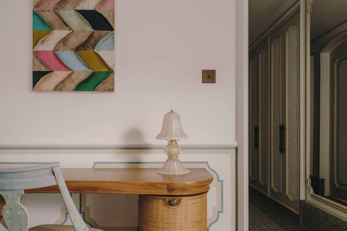 A room with a wooden desk, a wicker storage chest, a chair, and a colorful abstract painting on the wall.