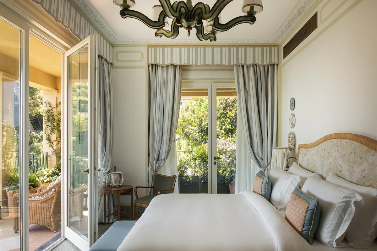 A hotel room with a large bed, a chair, a small table, and a view of the garden through the glass door.