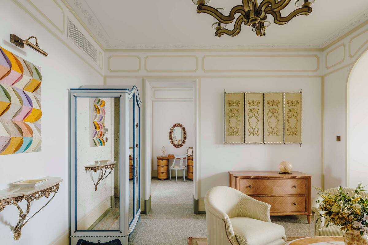 A room with a white and gold color scheme, featuring a mirror, a dresser, a chair, and a decorative wall panel.