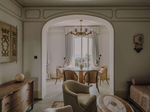 A dining area with a round table, chairs, and a chandelier. There is a wooden dresser and a sofa with patterned cushions.
