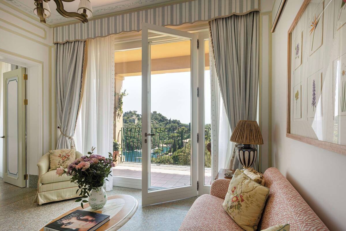A room with a sofa, a chair, a table with a vase and a book, and a view through a glass door to an outdoor area with a railing.