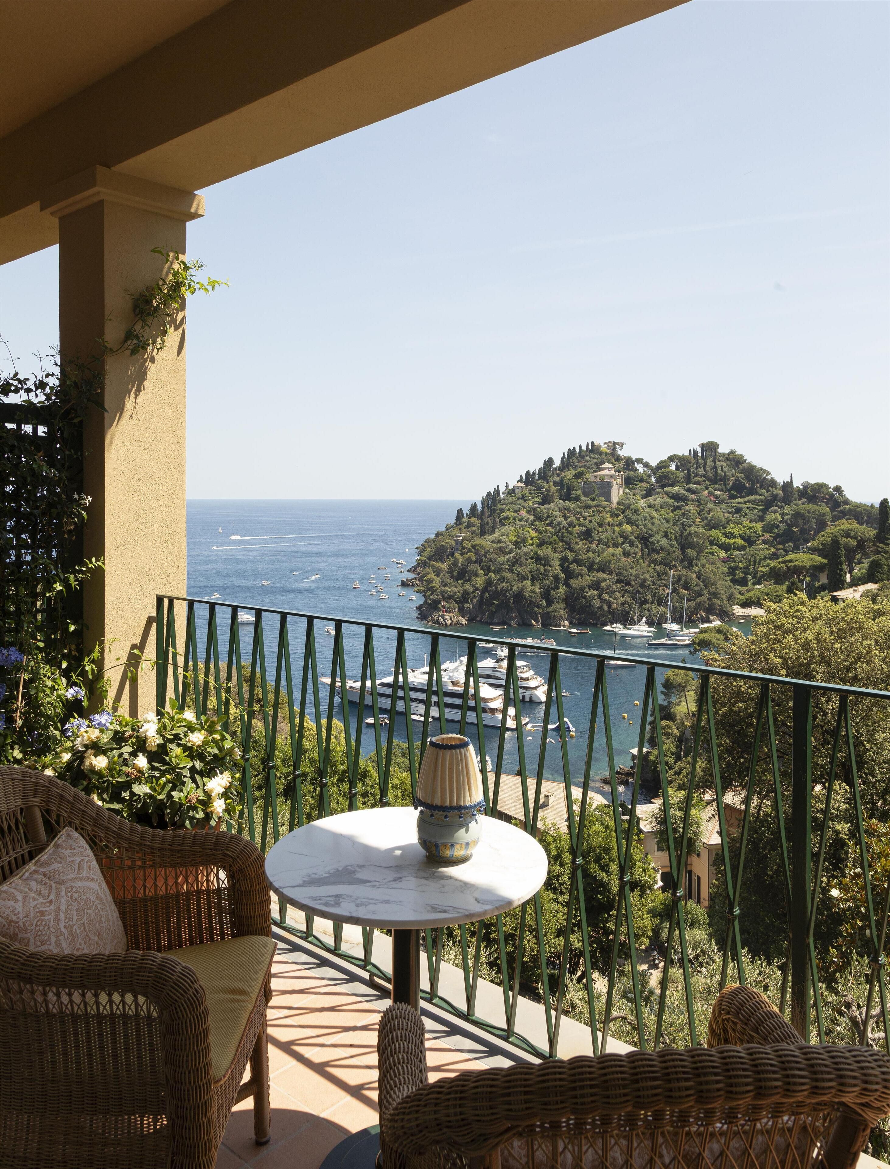 A balcony with wicker furniture, a table, and a view of the sea and a hill with buildings.