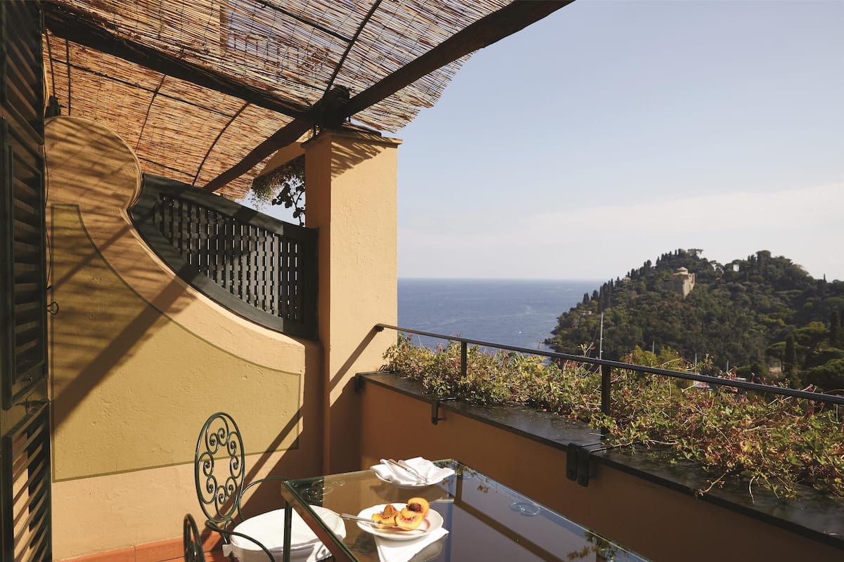 A balcony with a glass table, a chair, and a plate with food. There is a view of a hill with trees and a building in the distance.