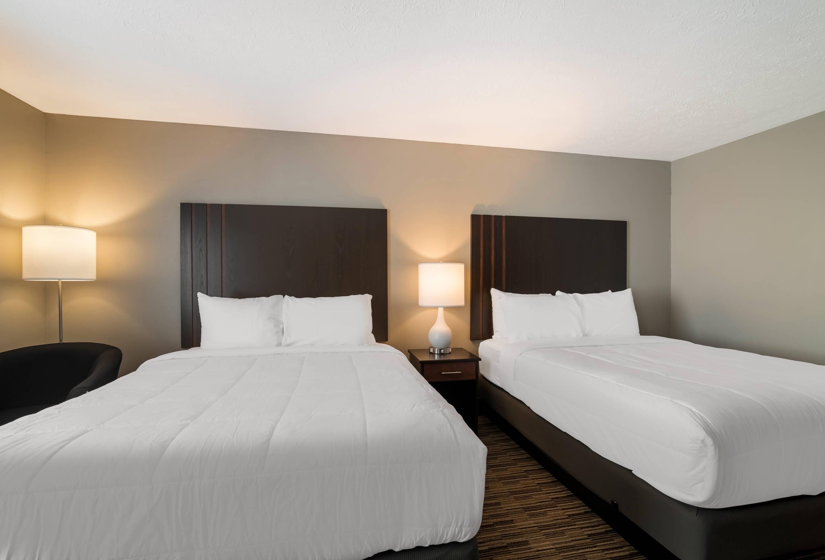 A hotel room with two beds, a nightstand with a lamp, and a chair.