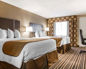 Two beds with white linens and brown headboards, a large window, and a desk with a chair and lamp.