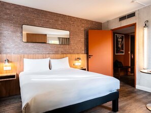 A hotel room with a brick wall, a bed with white linens, two bedside lamps, and a door leading to a bathroom. - ibis Styles Lyon Villeurbanne Parc de la Tête d'Or (Villeurbanne)