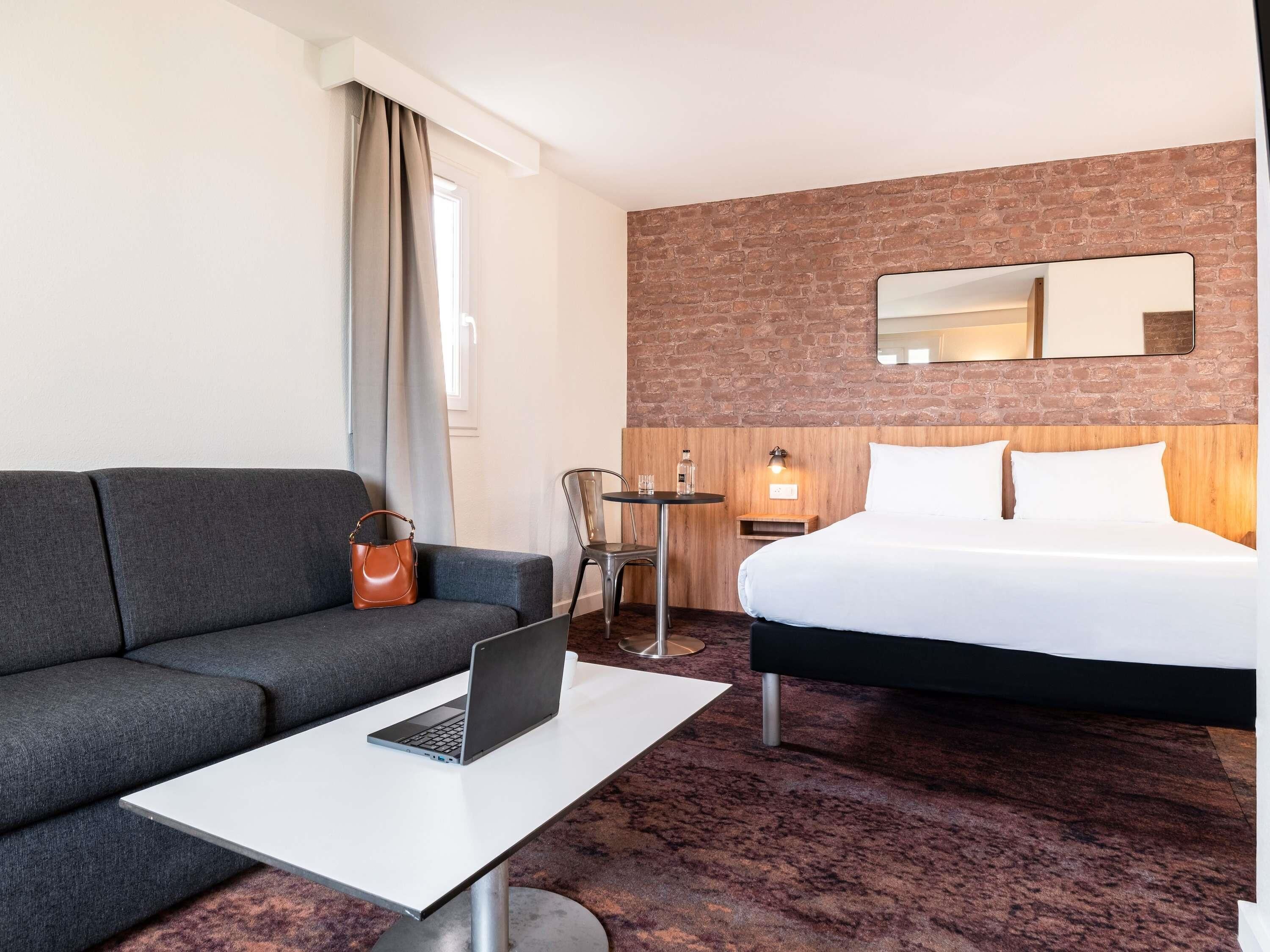 A hotel room with a grey sofa, a bed with white linens, a laptop on a small table, and a brick wall.