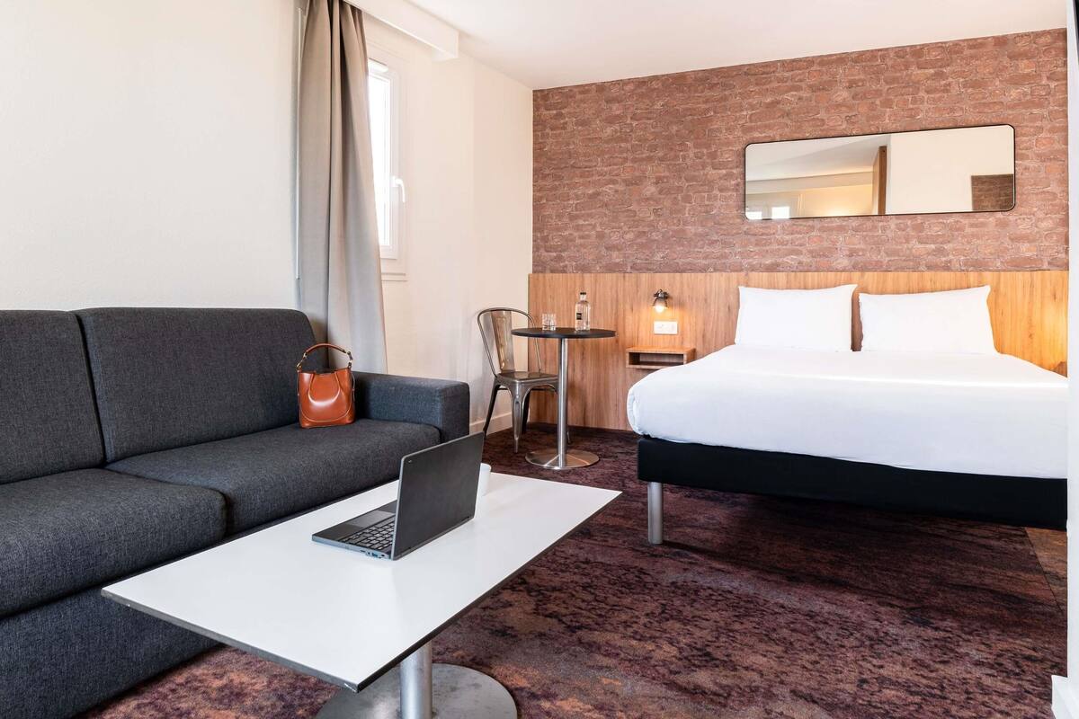 A hotel room with a grey sofa, a bed with white linens, a laptop on a small table, and a brick wall.