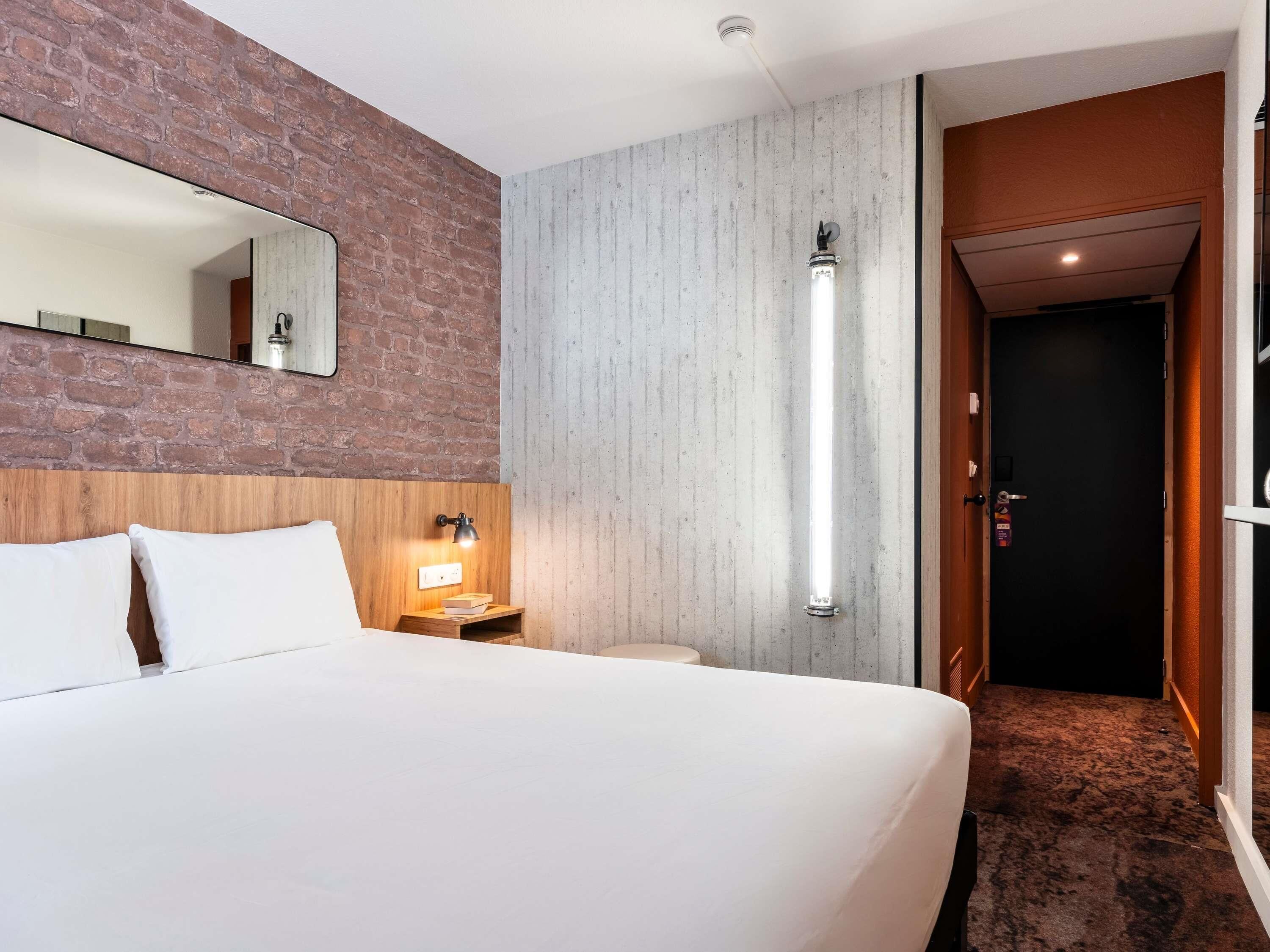 Habitación de hotel con una cama grande, cabecera de madera, mesita de noche con lámpara, pared de ladrillo decorativa, pared de concreto, lámpara vertical alta y puerta que da a otra habitación.