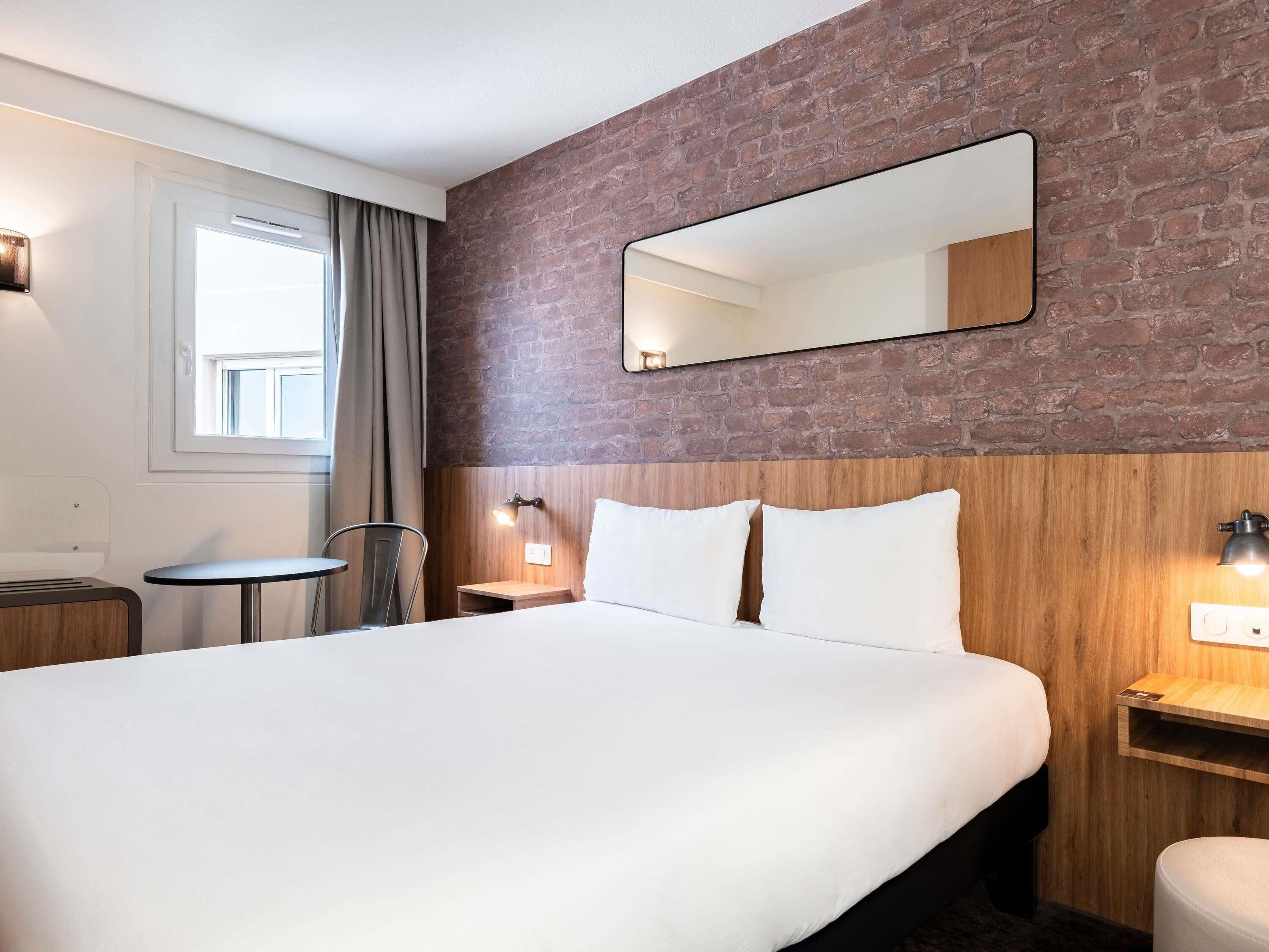 A hotel room with a large bed, a wooden headboard, a mirror, a small table, and a chair.