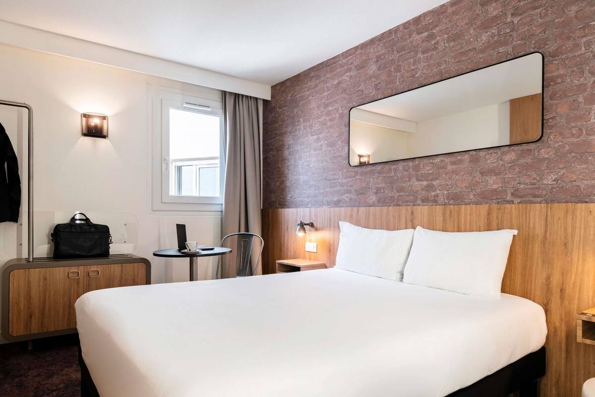 A hotel room with a large bed, a wooden headboard, a mirror, a small table with a chair, a desk, and a wall-mounted lamp.